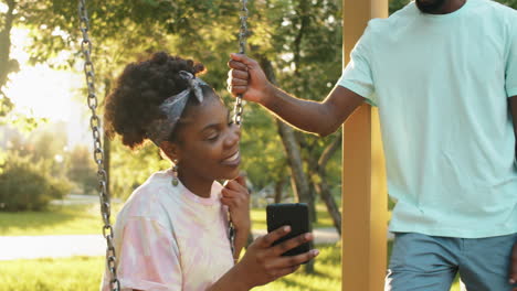 African-American-Friends-Chatting,-Using-Smartphone-and-Swinging-on-Swing-in-Park